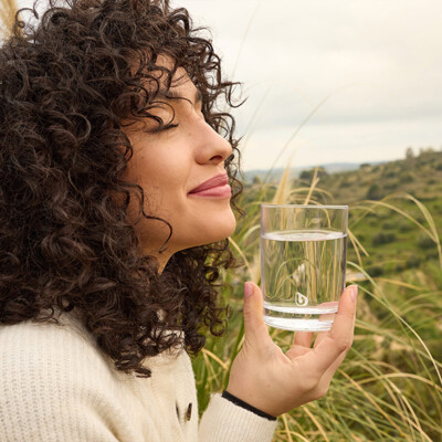 Eine lächelnde Frau mit lockigem Haar hält ein Glas klares Wasser und schließt entspannt die Augen.