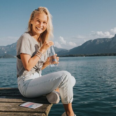 Hanna Hofinger sitzt entspannt auf einem Holzsteg am klaren Seeufer, genießt ein erfrischendes Wasser und lächelt in die Kamera. 