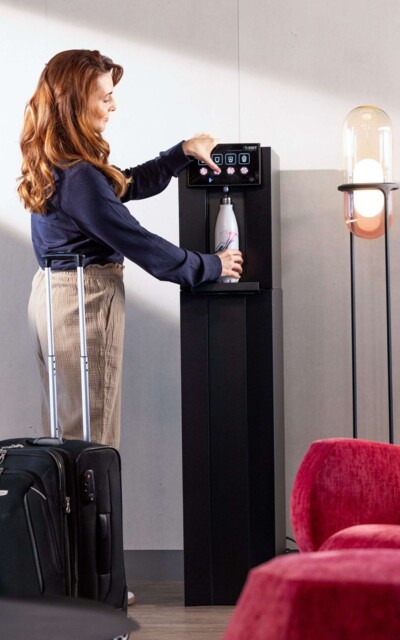 Eine Frau mit Rollkoffer befüllt ihre Flasche an einem BWT Wasserspender in einer modernen Hotellobby. 