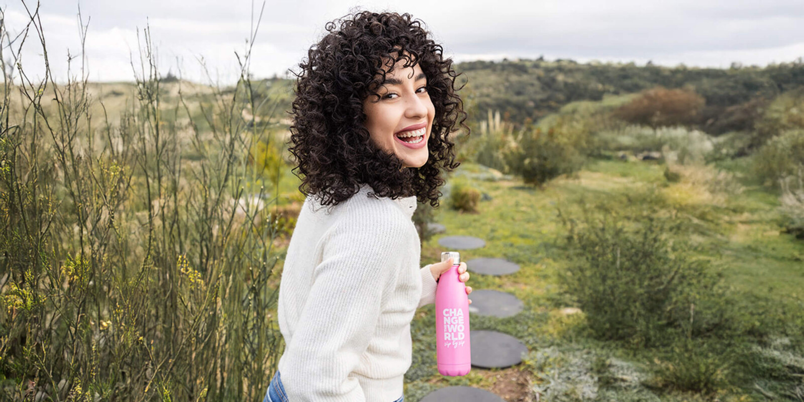Eine junge Frau mit lockigem Haar und weißem Pullover hält eine pinke Trinkflasche in der Hand.